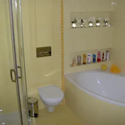 Jasna łazienka z narożną wanną, kabiną prysznicową, toaletą i półkami w ścianie z dekoracjami i kosmetykami, wykończona żółtymi płytkami.